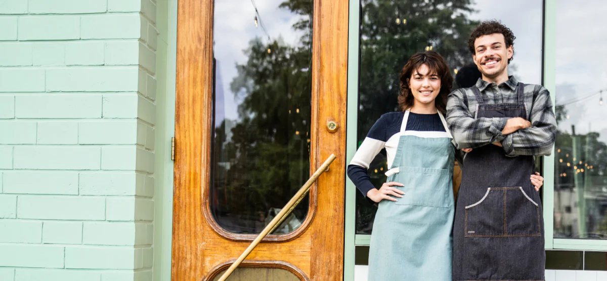 Two smiling coworkers wearing aprons stand outside a storefront with a wooden door and large window.